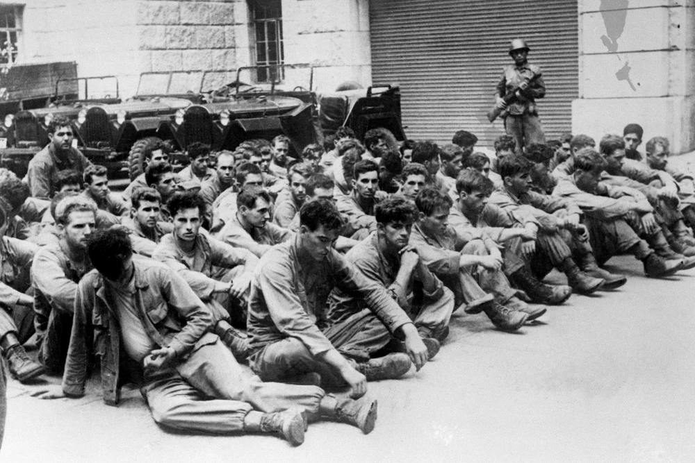 A group of uniformed men sit on the ground outside a utilitarian looking building, with armed guards standing around them. There are several military cars in the background.